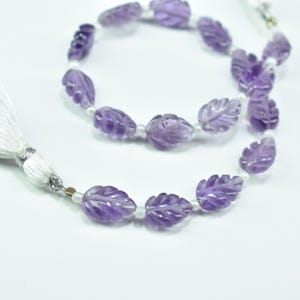 May include: A strand of amethyst beads carved in a leaf design. The purple beads are strung with small white beads. A white tassel with silver accents is attached to the end of the strand. The beads are displayed on a white background.