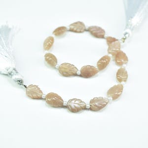 Op de afbeelding: Ketting met perzik-kleurige, bladvormige kralen met gesneden details, afgewisseld met kleine witte kralen. De ketting wordt gepresenteerd op een witte ondergrond, met een zilveren kwast aan de linkerkant.