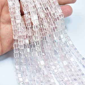 Può includere: Fili di perline di pietre preziose trasparenti, tagliate a forma quadrata. Le perline traslucide hanno una leggera sfumatura rosa e sono infilate su un filo sottile rosa. Le perline sono tenute in mano su sfondo bianco.