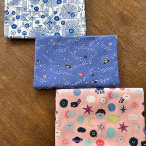 May include: Three fabric squares with different patterns. The top square is white with a blue geometric pattern. The middle square is blue with a white star pattern. The bottom square is pink with a white cloud and star pattern.