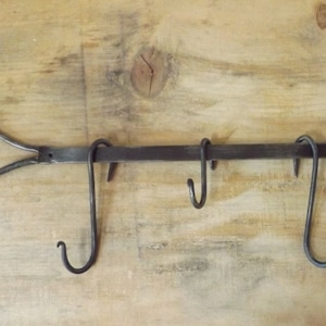 以下が含まれることがあります： 5つの湾曲したフックが付いた、手作りの鍛鉄製の壁掛けラック。ラックの両端にはハート型のデザインがあります。濃い色の金属製のラックは、明るい色の木製の表面に取り付けられています。