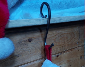 Puede incluir: Un soporte para calcetín navideño de metal forjado a mano en color gris oscuro, con un diseño en forma de S y un gancho superior curvado. El soporte se coloca sobre un borde de madera y sostiene un calcetín de felpa rojo y blanco.