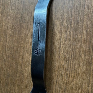 May include: A black, wrought iron door pull with an arrow design. The handle has a curved shape and features a decorative arrow etched into the center. The ends of the handle have a flared design with a single hole for mounting.