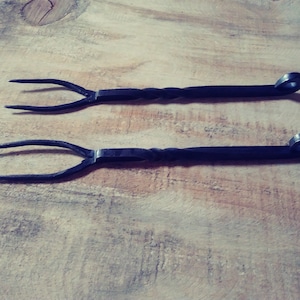 May include: Two black, hand-forged metal forks with a twisted handle design and a loop at the end. The forks have three tines each. The items are displayed on a light brown wooden surface.
