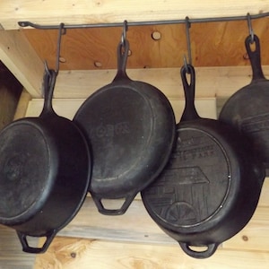 May include: Four black cast iron skillets hanging from a metal rack. The skillets are different sizes and shapes, with visible handles and some with embossed designs. The skillets are suspended from a metal bar attached to a wooden structure.