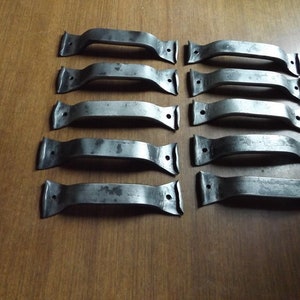 May include: Ten weathered, metal drawer pulls arranged on a wooden surface. Each pull has a curved handle with a rectangular base and two screw holes. The metal has a dark, aged appearance.