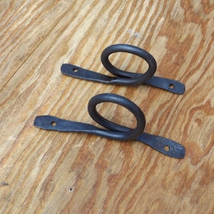 May include: Two handcrafted, black metal ring pulls. Each pull features a circular ring attached to a flat, rectangular base with a single mounting hole. The metal has a textured, hammered appearance, suggesting a rustic, artisanal style.