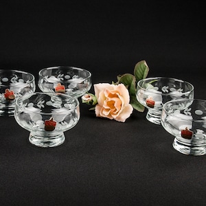 Könnte beinhalten: Fünf klare Glasschalen für Desserts mit einem geätzten Blumenmuster. Die Schalen haben einen Sockel und stehen auf einer schwarzen Oberfläche.