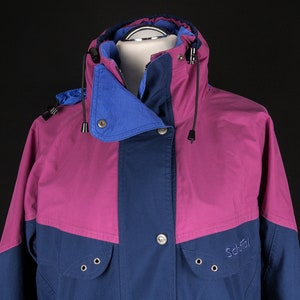 Könnte beinhalten: Eine blaue und rosafarbene Jacke mit Kapuze und Reißverschluss. Die Jacke hat ein Schöffel-Logo auf der Vorderseite.