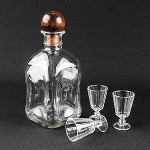 Könnte beinhalten: Eine klare Glaskaraffe mit einem Holzstopfen und zwei klaren Schnapsgläsern. Die Karaffe hat ein einzigartiges, strukturiertes Design.