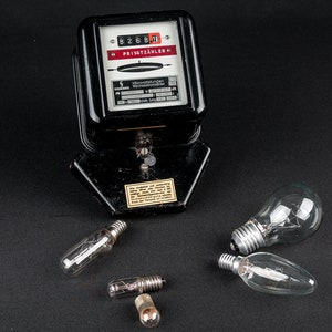 Könnte beinhalten: Ein schwarzer Stromzähler mit einer digitalen Anzeige, die "8268.1" anzeigt, und dem Text "Privatzähler". Vier Glühbirnen liegen verstreut um den Zähler herum.