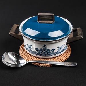 Könnte beinhalten: Ein weiß-blauer Emaille-Kochtopf mit floralem Muster, blauem Deckel und Holzgriff steht auf einem gewebten Untersetzer. Ein silberner Löffel liegt vor dem Topf. Der Topf hat einen Durchmesser von etwa 20 cm.