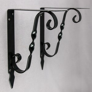 Könnte beinhalten: Zwei schwarze Metall-Regalhalterungen mit einem dekorativen Schnörkeldesign. Die Halterungen sind an einer Wand befestigt.