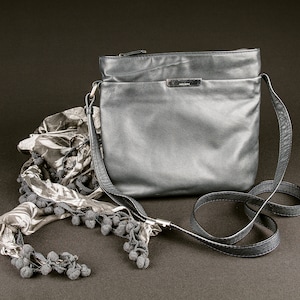Könnte beinhalten: Eine silberne Leder-Umhängetasche mit einem schwarzen Riemen und einem schwarz-weißen gemusterten Schal mit Bommeln.