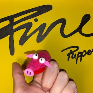 Könnte beinhalten: Eine rosa Schweine-Fingerpuppe mit weißen Wackelaugen und rosa Ohren und Schnauze. Die Puppe wird vor einem gelben Hintergrund mit den Worten "Fine Puppets" in Schwarz gehalten.
