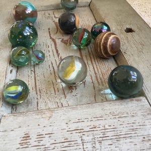 Puede incluir: Una colección de canicas de vidrio en varios colores y patrones, incluyendo verde, azul, amarillo y marrón. Las canicas se exhiben sobre una superficie de madera clara y desgastada, mostrando sus cualidades reflectantes y diseños intrincados. Algunas canicas tienen remolinos y rayas.
