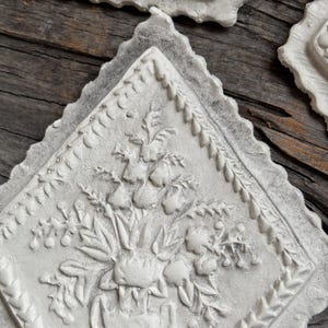 Op de afbeelding: Close-up van verschillende witte, ruitvormige decoratieve plaquettes met bloemreliëf. Elke plaquette heeft een centrale vaas met bloemen, omgeven door een gedetailleerde rand. De plaquettes zijn geplaatst op een rustiek, verweerd houten oppervlak.