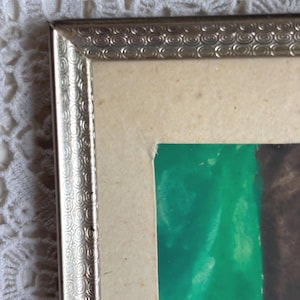 May include: Close-up of a framed artwork. The frame is silver-coloured with a decorative pattern. The artwork features green and brown colours. The paper has a light beige colour.