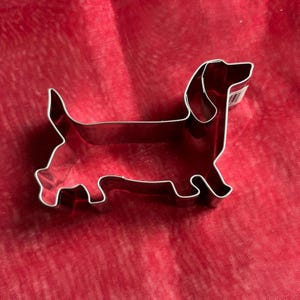 Könnte beinhalten: Glänzender, silberfarbener Metall-Ausstecher in Form eines Dackelhundes. Der Ausstecher befindet sich auf rotem Hintergrund. Der Hund ist im Seitenprofil dargestellt, mit seinem langen Körper und kurzen Beinen.