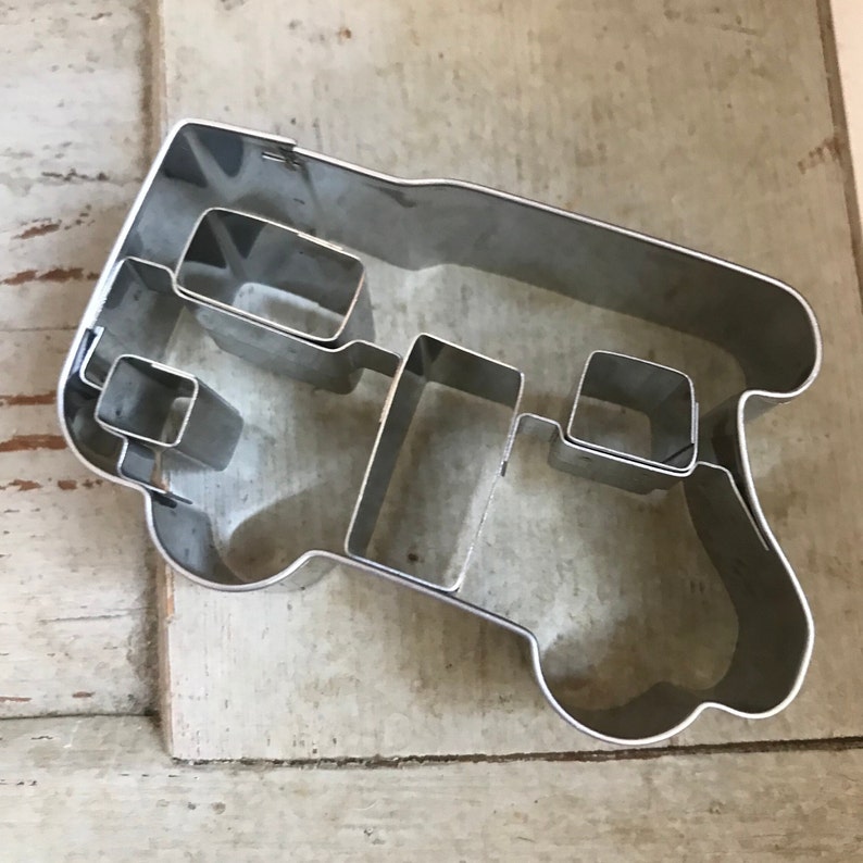 May include: A silver-colored metal cookie cutter shaped like a camper van. The cutter features a continuous outer frame with internal metal dividers that create distinct sections for the windows, door, and wheels of the vehicle design.