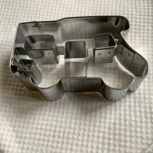 May include: A silver-colored metal cookie cutter shaped like a tractor. The cutter features a distinct outline of a tractor body with internal metal dividers that create separate sections for the wheels, cabin, and engine hood areas.