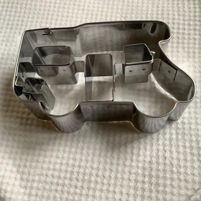 May include: A silver-colored metal cookie cutter shaped like a tractor. The cutter features a distinct outline of a tractor body with internal metal dividers that create separate sections for the wheels, cabin, and engine hood areas.
