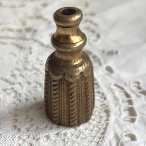 Könnte beinhalten: Ein kleiner, antiker Flaschenverschluss aus Messing. Der Verschluss hat ein dekoratives, strukturiertes Design am unteren Teil und eine abgerundete Oberseite. Das Messing hat eine warme, gealterte Patina, die auf seinen Vintage-Charakter hindeutet.