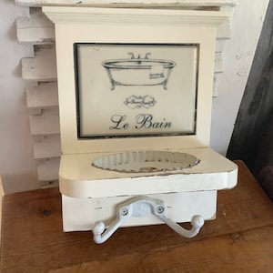 Könnte beinhalten: Cremefarbene, rustikale Holz-Wanddekoration mit einem gerahmten Bild einer Vintage-Badewanne, den Worten "Le Bain" und zwei Haken. Die Dekoration hat ein kleines Regal und ist für den Badezimmergebrauch konzipiert.