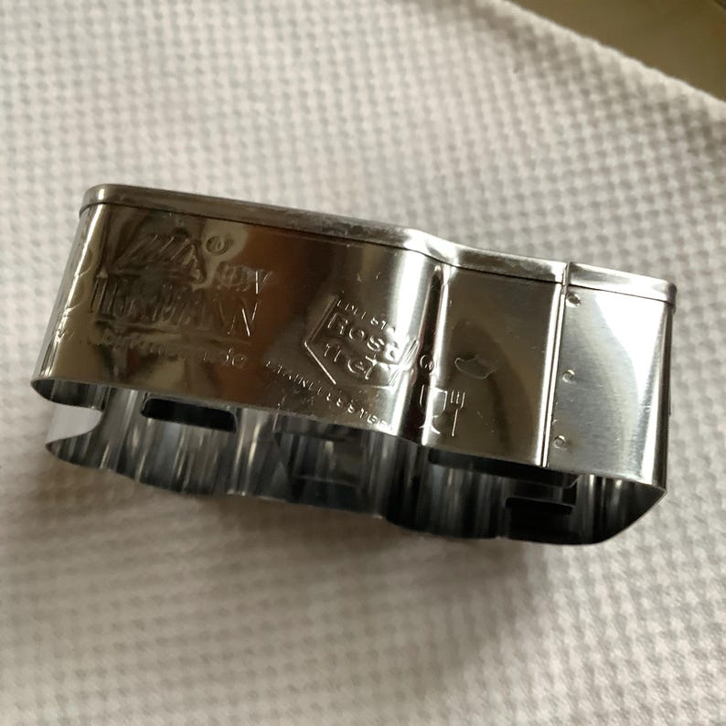 May include: A silver-colored stainless steel cookie cutter with a reflective surface. The side of the cutter features engraved text including the brand name Birkmann, the word Rostfrei, and a food-safe symbol. The metal is shaped into a curved form.