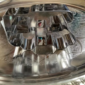 May include: A silver-colored metal cookie cutter shaped like a vehicle, featuring a rectangular cabin and a rounded front section. The cutter has a reflective, polished surface and is constructed from a single continuous strip of bent metal.