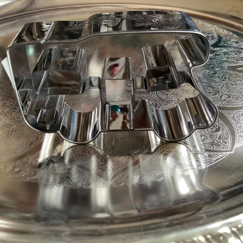 May include: A silver-colored metal cookie cutter shaped like a vehicle, featuring a rectangular cabin and a rounded front section. The cutter has a reflective, polished surface and is constructed from a single continuous strip of bent metal.