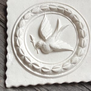Könnte beinhalten: Eine quadratische, weiße Reliefskulptur mit einer Taube, die einen Olivenzweig in einem kreisförmigen Kranz trägt. Die Taube ist im Flug dargestellt, und der Kranz besteht aus sich wiederholenden Blattformen. Die Kanten der Skulptur sind rau.