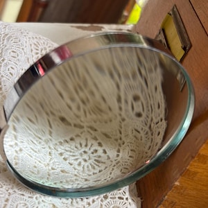 Könnte beinhalten: Eine runde Lupe mit einem polierten, silberfarbenen Metallrahmen und einem schwarzen Griff. Die kreisförmige Glaslinse reflektiert ein weißes Spitzenstoffmuster, und das Gerät befindet sich neben einem hellbraunen Holzobjekt mit einem Metallschildhalter.