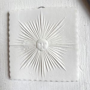 May include: A white, square relief carving of a smiling sun with radiating beams. The sun has a face with closed eyes and a smiling mouth. The edges of the carving are uneven, giving it a rustic appearance. A small loop is attached at the top for hanging.