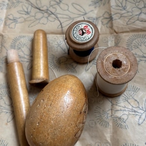 Könnte beinhalten: Eine Sammlung von Vintage-Nähwerkzeugen, darunter Holzspulen mit Faden, ein eiförmiger Stopfpilz aus Holz und Holzgriffe. Die Artikel sind auf einem Hintergrund aus Papier mit floralem Muster angeordnet.