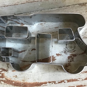 May include: A silver-colored metal cookie cutter shaped like a camper van. The outline forms the exterior of the vehicle, while internal metal strips create the shapes of windows and doors within the main frame of the cookie cutter.