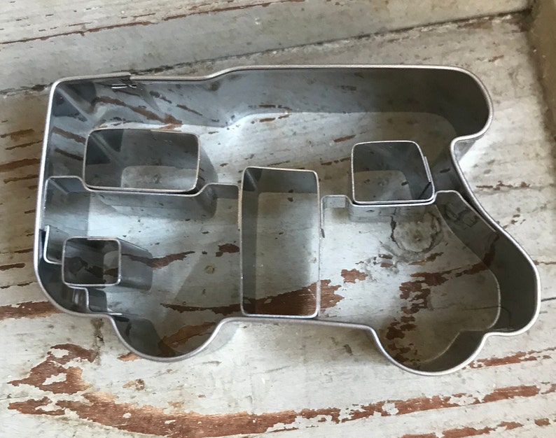 May include: A silver-colored metal cookie cutter shaped like a camper van. The outline forms the exterior of the vehicle, while internal metal strips create the shapes of windows and doors within the main frame of the cookie cutter.