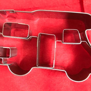 May include: A silver-colored metal cookie cutter shaped like a camper van. The outline forms the exterior of the vehicle, while internal metal strips create the shapes of windows and a door. The cookie cutter has a shiny, reflective finish.