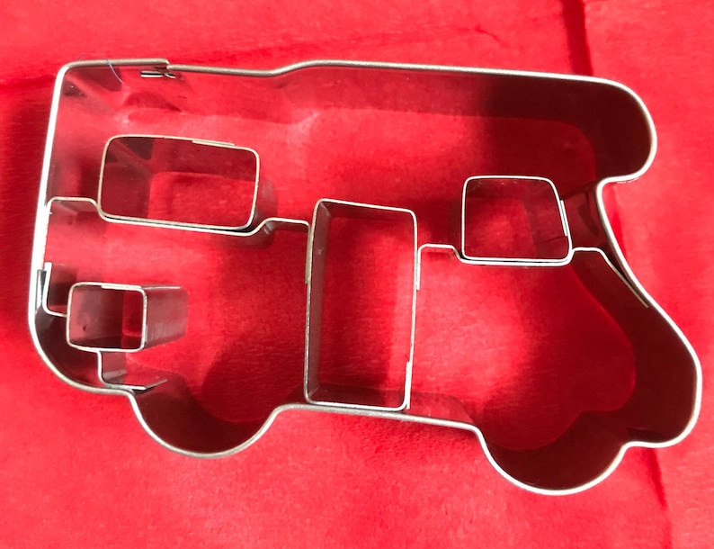 May include: A silver-colored metal cookie cutter shaped like a camper van. The outline forms the exterior of the vehicle, while internal metal strips create the shapes of windows and a door. The cookie cutter has a shiny, reflective finish.