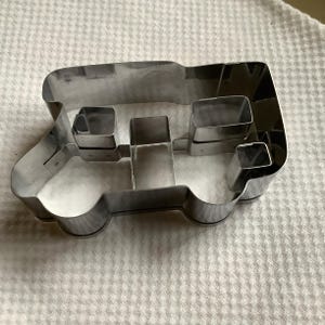 May include: A silver-colored metal cookie cutter shaped like a camper van. The cutter features a rectangular outline with internal dividers that create distinct sections for the windows and body panels of the vehicle design.