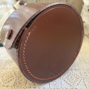 Könnte beinhalten: Eine braune Ledertasche mit runder Form. Die Tasche hat eine glänzende Oberfläche und ist mit orangefarbenem Faden an den Rändern genäht. Ein kleiner, brauner Knopf sichert den Deckel.