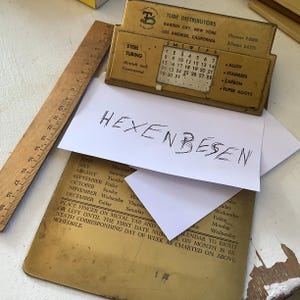 Könnte beinhalten: Ein Nahaufnahme eines Messingkalenders mit einer weißen Karte darauf. Auf der Karte steht der deutsche Begriff "Hexenbesen". Der Kalender zeigt eine Liste der Monate und Wochentage sowie eine kleine Metalllasche, die verschoben werden kann, um das aktuelle Datum anzuzeigen.
