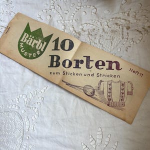 May include: A vintage knitting pattern booklet, tan in colour, with the text "Bärbl Muster" in a green emblem. The booklet features the words "10 Borten" and a drawing of a sweater and yarn. It rests on a white lace surface.