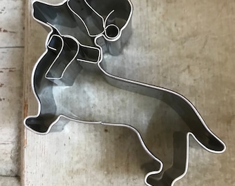 May include: A silver metal cookie cutter shaped like a dachshund wearing a Santa hat. The cookie cutter is designed to cut out a Christmas-themed treat.