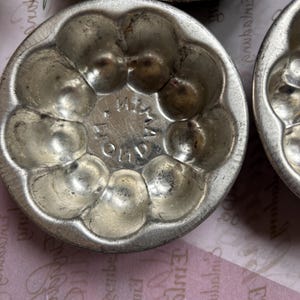 Könnte beinhalten: Nahaufnahme mehrerer antiker, silberfarbener Metallformen, jede mit einer kreisförmigen Form und mehreren abgerundeten Hohlräumen. Die Formen haben ein Vintage-Aussehen, was auf Alter und Gebrauch hindeutet. Das Wort "IMA" ist auf einer Form sichtbar.