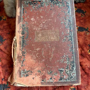 May include: An antique, weathered hardback book with a faded red cover featuring black decorative floral borders and a central rectangular label with the text Kochbuch. The spine is frayed and partially detached, showing signs of significant age.