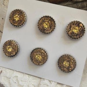 May include: A set of six circular buttons arranged on a white card. Each button features a textured, metallic bronze-coloured centre with a serrated silver-toned rim and a single, small, faceted yellow crystal embedded in the centre of each piece.