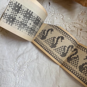 Op de afbeelding: Open boek met pagina's met zwart-witte kruissteekpatronen. Eén pagina toont een geometrisch ontwerp, terwijl een andere het woord "SWAN" in een herhalend patroon bevat. De pagina's zijn verouderd met een gelige tint, wat wijst op vintage of antieke oorsprong.