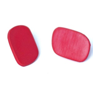 Puede incluir: Un par de pendientes ovalados de color rojo. Los pendientes están hechos de un material liso y mate.
