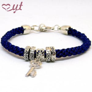 Könnte beinhalten: Ein blaues geflochtenes Armband mit silbernen Glücksbringerperlen, darunter eine Krone und ein Schlüsselanhänger.